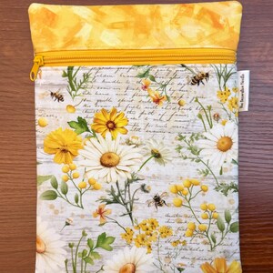 K&ouml;nnte beinhalten: Ein rechteckiger Stoffbeutel mit gelbem Rei&szlig;verschluss und einem oberen Rand mit gelbem abstraktem Muster. Der Hauptteil zeigt einen Druck aus wei&szlig;en G&auml;nsebl&uuml;mchen, gelben Wildblumen und Bienen auf einem Hintergrund aus kursiver Schrift und Holzmaserung.