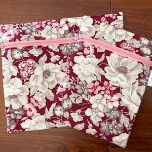 May include: Two rectangular zippered pouches featuring a floral pattern with large cream and light gray flowers on a deep burgundy background. Each pouch has a light pink zipper closure across the top and a small white fabric tag on the side.