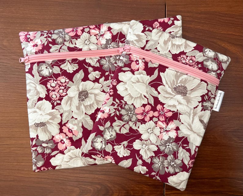 May include: Two rectangular zippered pouches featuring a floral pattern with large cream and light gray flowers on a deep burgundy background. Each pouch has a light pink zipper closure across the top and a small white fabric tag on the side.