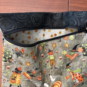 May include: A Halloween-themed zippered pouch with a black zipper and a black patterned top. The pouch features a gray background with whimsical illustrations of witches, pumpkins, stars, and cats. The interior lining has a pumpkin pattern.