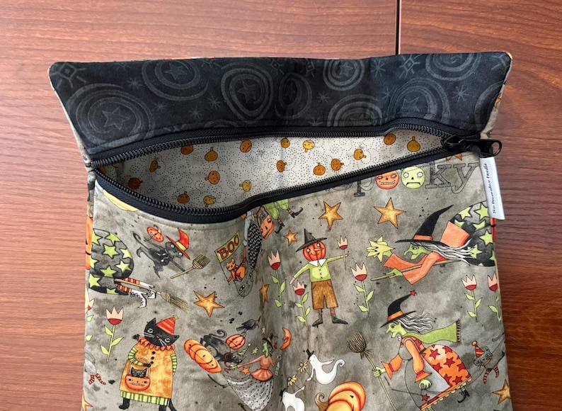 May include: A Halloween-themed zippered pouch with a black zipper and a black patterned top. The pouch features a gray background with whimsical illustrations of witches, pumpkins, stars, and cats. The interior lining has a pumpkin pattern.