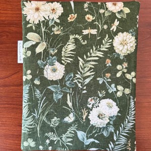 Pu&ograve; includere: Una copertina per libri in tessuto verde con un motivo floreale. Il design presenta fiori bianchi, felci e farfalle su uno sfondo verde scuro. Il testo "the decorative beetle" &egrave; su una piccola etichetta bianca.