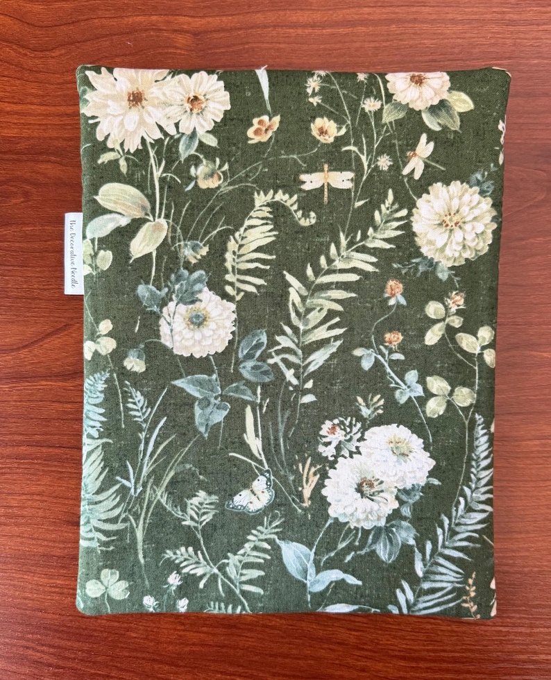 Pu&ograve; includere: Una copertina per libri in tessuto verde con un motivo floreale. Il design presenta fiori bianchi, felci e farfalle su uno sfondo verde scuro. Il testo "the decorative beetle" &egrave; su una piccola etichetta bianca.