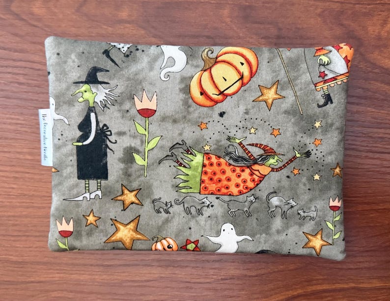 May include: A rectangular, gray Halloween-themed pouch featuring illustrations of witches, pumpkins, stars, cats, and ghosts. The design includes a witch in a black dress and hat, and another flying with a wand. The pouch has a small white tag on the side.