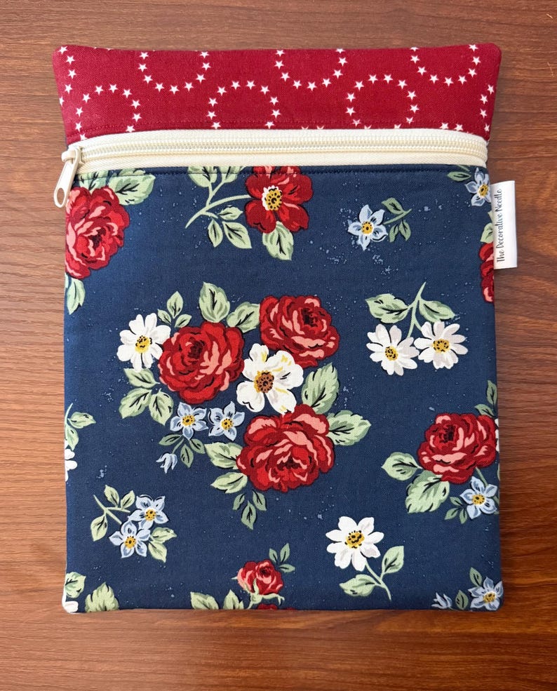 K&ouml;nnte beinhalten: Ein rechteckiger Rei&szlig;verschlussbeutel mit floralem Muster. Der Hauptteil ist marineblau mit roten Rosen, wei&szlig;en G&auml;nsebl&uuml;mchen und kleinen blauen Blumen. Der obere Teil ist rot mit wei&szlig;en Sternformen. Ein cremefarbener Rei&szlig;verschluss ist sichtbar.
