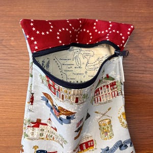 Op de afbeelding: Een stoffen etui met een rood-wit sterrenpatroon op de bovenklep. De hoofdband heeft een lichtblauwe achtergrond met historische illustraties en een kaart. Een zwarte ritssluiting sluit de opening af.