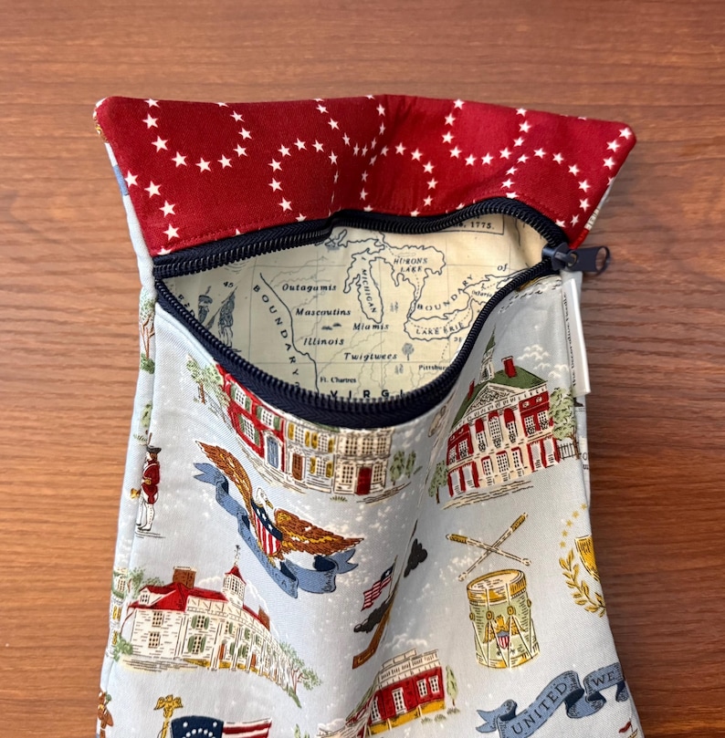 Op de afbeelding: Een stoffen etui met een rood-wit sterrenpatroon op de bovenklep. De hoofdband heeft een lichtblauwe achtergrond met historische illustraties en een kaart. Een zwarte ritssluiting sluit de opening af.