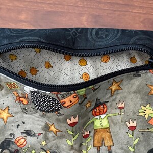 May include: A zippered pouch with a Halloween-themed pattern. The exterior features cartoon illustrations of pumpkins, stars, and characters in a muted color palette. The interior lining is a light gray with small orange pumpkin designs. The zipper is black.