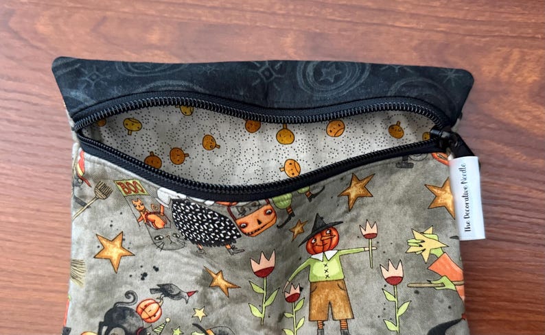 May include: A zippered pouch with a Halloween-themed pattern. The exterior features cartoon illustrations of pumpkins, stars, and characters in a muted color palette. The interior lining is a light gray with small orange pumpkin designs. The zipper is black.