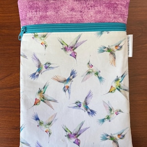 May include: A rectangular pouch with a white background, featuring a pattern of colorful hummingbirds in flight. The top section is a textured purple fabric, and a teal zipper secures the opening. The label reads "The Decorative Needle".