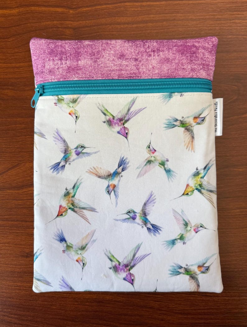 May include: A rectangular pouch with a white background, featuring a pattern of colorful hummingbirds in flight. The top section is a textured purple fabric, and a teal zipper secures the opening. The label reads "The Decorative Needle".