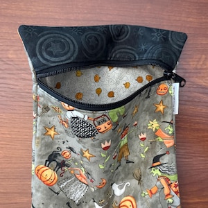 May include: A zippered pouch with a Halloween-themed pattern. The pouch features a black zipper and a gray interior with orange pumpkins and other Halloween characters. The exterior fabric has a gray background with Halloween figures, stars, and pumpkins.