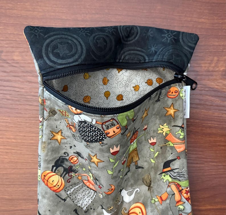 May include: A zippered pouch with a Halloween-themed pattern. The pouch features a black zipper and a gray interior with orange pumpkins and other Halloween characters. The exterior fabric has a gray background with Halloween figures, stars, and pumpkins.