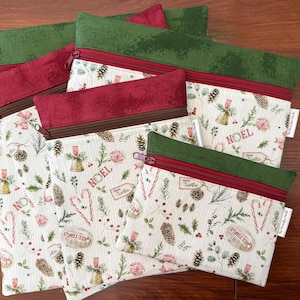 May include: A set of three fabric zipper pouches in varying sizes featuring a cream background with a Christmas-themed print of pinecones, candy canes, bells, and the text NOEL. Each pouch has a dark red zipper and a solid green fabric top border.