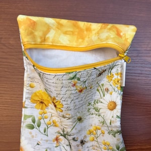 K&ouml;nnte beinhalten: Ein Stoffbeutel mit Rei&szlig;verschluss, gelbem oberen Rand und wei&szlig;em Grund, bedruckt mit einem Muster aus gelben G&auml;nsebl&uuml;mchen, Wildblumen und kursivem Text. Der Beutel verf&uuml;gt &uuml;ber einen gelben Rei&szlig;verschluss und ein kleines wei&szlig;es Seitenetikett.