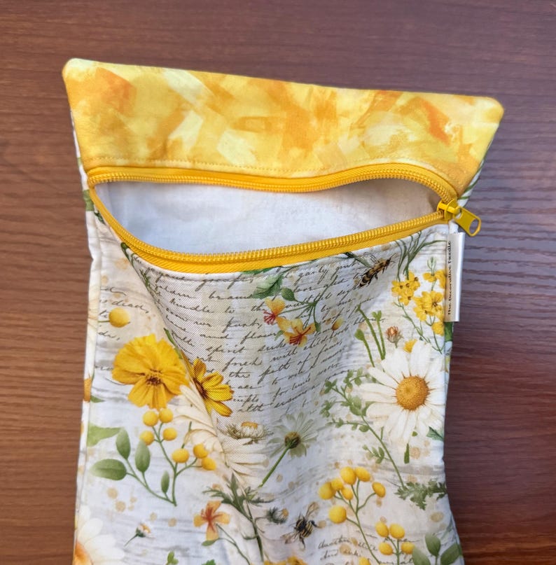 K&ouml;nnte beinhalten: Ein Stoffbeutel mit Rei&szlig;verschluss, gelbem oberen Rand und wei&szlig;em Grund, bedruckt mit einem Muster aus gelben G&auml;nsebl&uuml;mchen, Wildblumen und kursivem Text. Der Beutel verf&uuml;gt &uuml;ber einen gelben Rei&szlig;verschluss und ein kleines wei&szlig;es Seitenetikett.