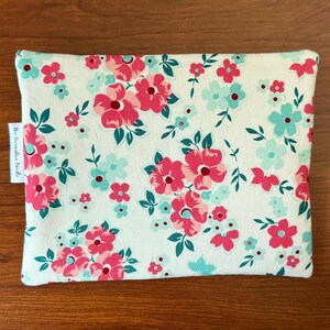 Puede incluir: Una bolsa rectangular con un estampado floral de flores rosas y turquesas y hojas verdes sobre un fondo crema. La bolsa tiene una etiqueta que dice "The Decorative Needle."
