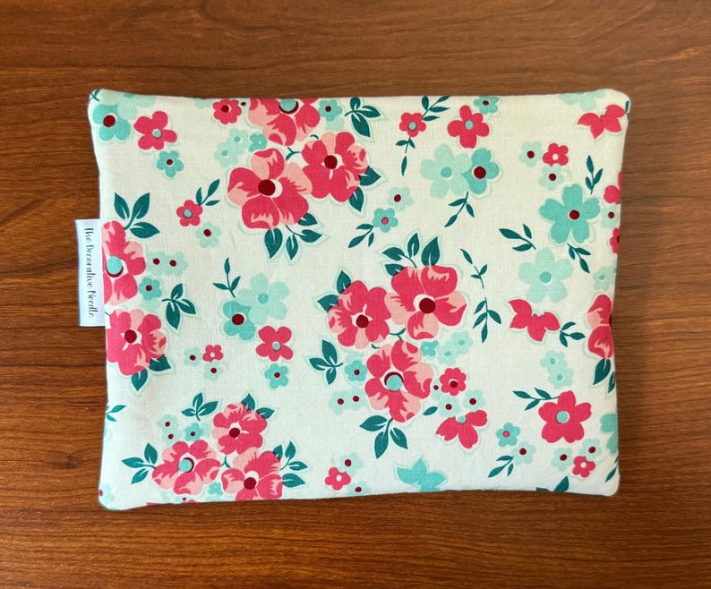Puede incluir: Una bolsa rectangular con un estampado floral de flores rosas y turquesas y hojas verdes sobre un fondo crema. La bolsa tiene una etiqueta que dice "The Decorative Needle."