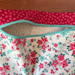 Puede incluir: Una bolsa con estampado floral con la parte superior roja y una cremallera turquesa. La bolsa presenta un fondo blanco con flores rosas y turquesas. La parte superior de la bolsa es roja con peque&ntilde;as flores blancas. El texto "The Dreamer Heads" est&aacute; en una etiqueta blanca.