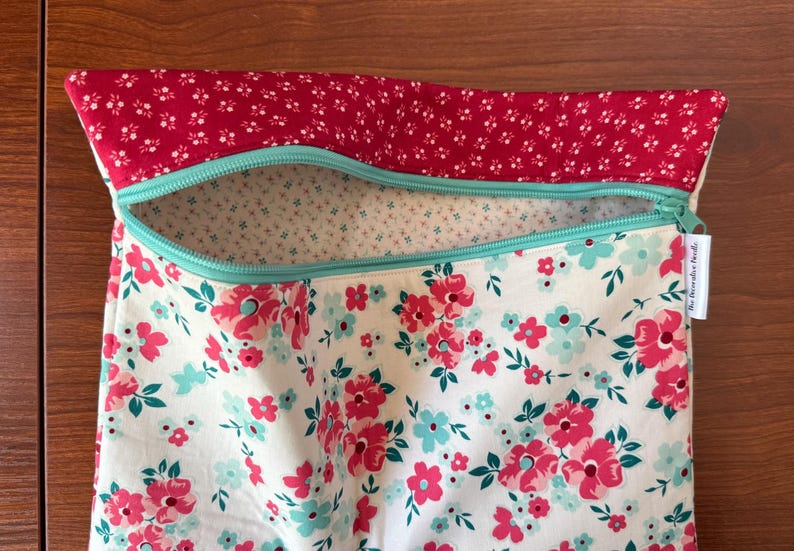 Puede incluir: Una bolsa con estampado floral con la parte superior roja y una cremallera turquesa. La bolsa presenta un fondo blanco con flores rosas y turquesas. La parte superior de la bolsa es roja con peque&ntilde;as flores blancas. El texto "The Dreamer Heads" est&aacute; en una etiqueta blanca.