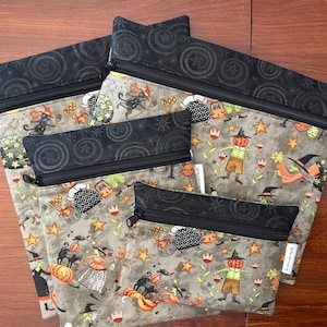 May include: A set of five Halloween-themed zippered pouches. The pouches feature a gray background with a repeating pattern of pumpkins, witches, and stars. The top and zipper are black. The pouches vary in size.