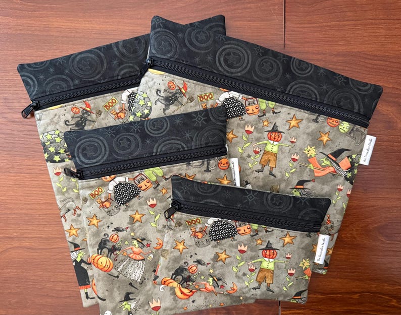 May include: A set of five Halloween-themed zippered pouches. The pouches feature a gray background with a repeating pattern of pumpkins, witches, and stars. The top and zipper are black. The pouches vary in size.