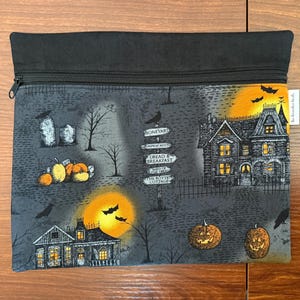 May include: A rectangular zippered pouch featuring a dark gray Halloween-themed fabric with a print of haunted houses, jack-o'-lanterns, bare trees, tombstones, bats, and a signpost with text including Boneyard, Dread & Breakfast, and The Rotten Cottage.