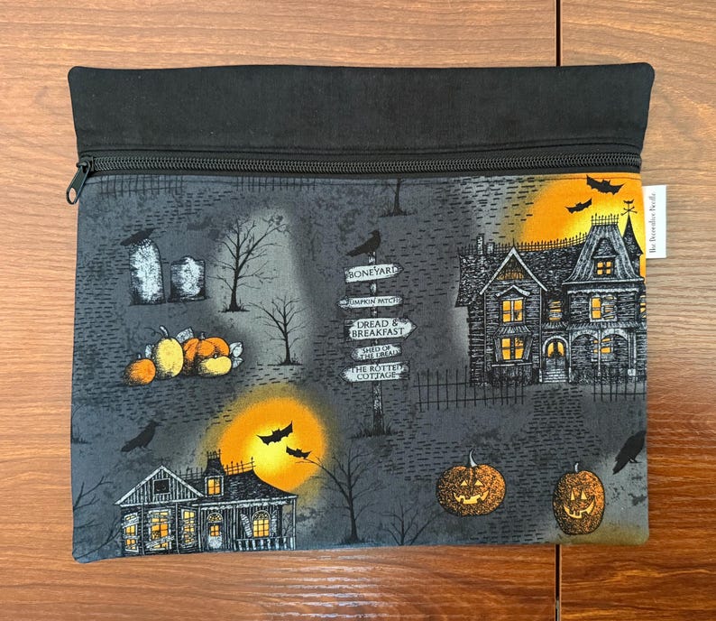 May include: A rectangular zippered pouch featuring a dark gray Halloween-themed fabric with a print of haunted houses, jack-o'-lanterns, bare trees, tombstones, bats, and a signpost with text including Boneyard, Dread & Breakfast, and The Rotten Cottage.