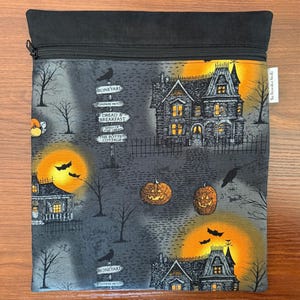 May include: A square fabric pouch with a black zipper closure featuring a Halloween print. The design includes haunted houses, glowing orange moons, bats, jack-o'-lanterns, and signposts labeled Boneyard, Dread & Breakfast, and The Rotted Cottage.