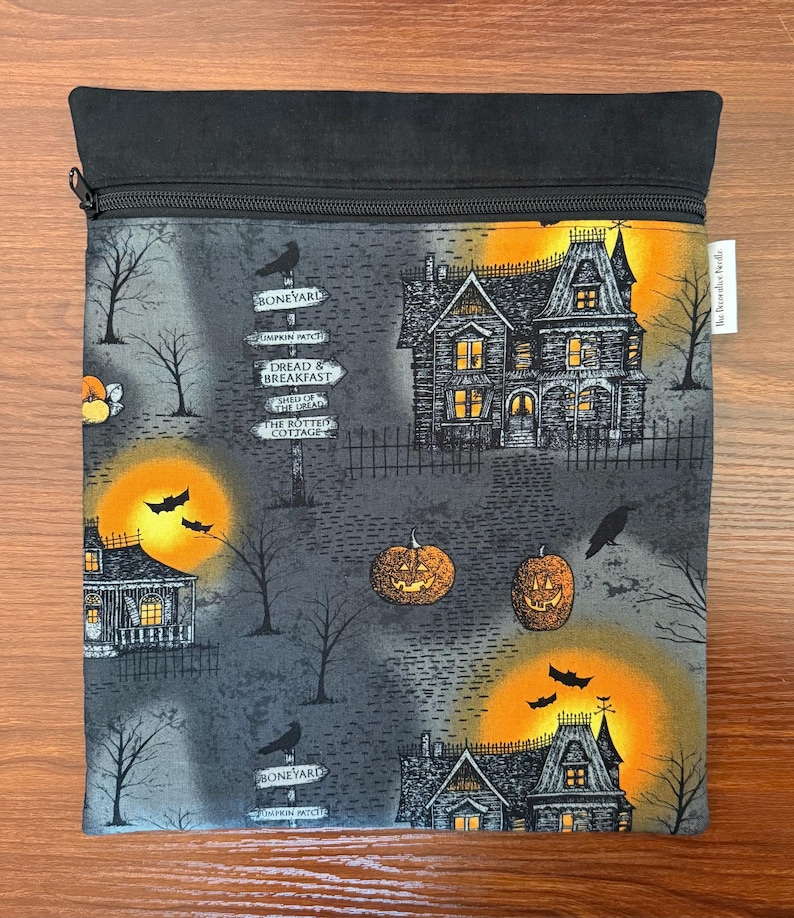 May include: A square fabric pouch with a black zipper closure featuring a Halloween print. The design includes haunted houses, glowing orange moons, bats, jack-o'-lanterns, and signposts labeled Boneyard, Dread & Breakfast, and The Rotted Cottage.
