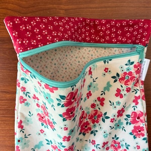 Puede incluir: Una bolsa de tela floral con la parte superior roja y cremallera turquesa. La bolsa presenta un fondo crema con flores rosas y turquesas. El forro interior es de tela crema con peque&ntilde;os estampados florales. La bolsa est&aacute; abierta.