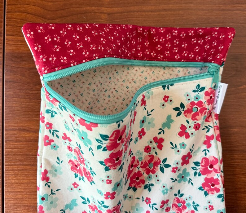 Puede incluir: Una bolsa de tela floral con la parte superior roja y cremallera turquesa. La bolsa presenta un fondo crema con flores rosas y turquesas. El forro interior es de tela crema con peque&ntilde;os estampados florales. La bolsa est&aacute; abierta.