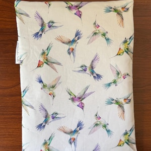May include: A rectangular, off-white pillowcase featuring a repeating pattern of colorful hummingbirds. The birds are depicted in flight with wings spread, showcasing shades of purple, blue, green, and orange. The pillowcase is set against a wooden surface.