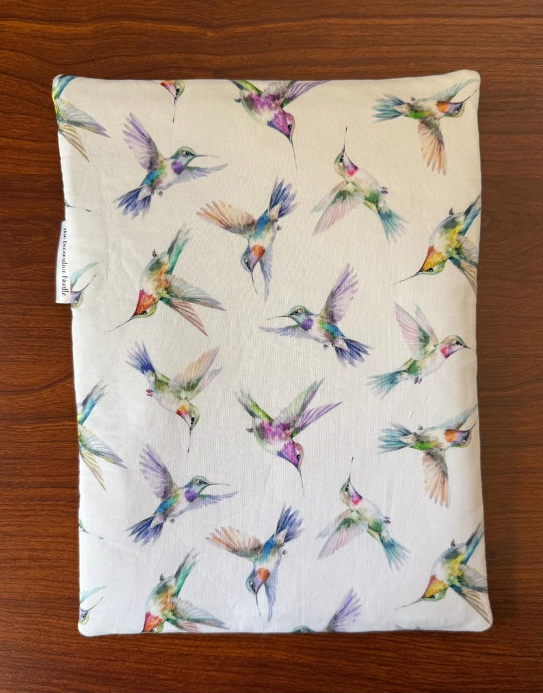 May include: A rectangular, off-white pillowcase featuring a repeating pattern of colorful hummingbirds. The birds are depicted in flight with wings spread, showcasing shades of purple, blue, green, and orange. The pillowcase is set against a wooden surface.