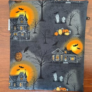 May include: A rectangular fabric pouch featuring a Halloween-themed print on a dark gray background. The pattern includes illustrations of haunted houses, glowing orange moons, black bats, bare trees, tombstones, and orange pumpkins.