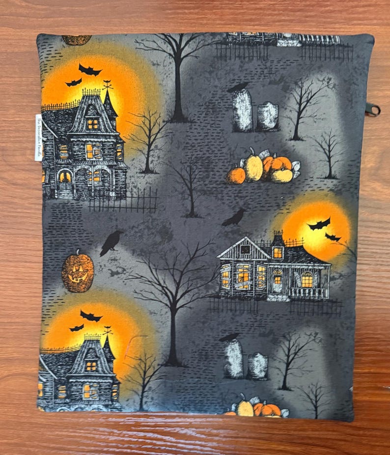 May include: A rectangular fabric pouch featuring a Halloween-themed print on a dark gray background. The pattern includes illustrations of haunted houses, glowing orange moons, black bats, bare trees, tombstones, and orange pumpkins.