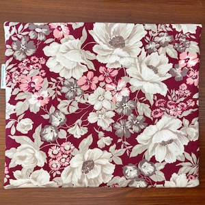 May include: A square pillow cover featuring a deep burgundy background with a dense floral pattern of large cream, light pink, and muted grey flowers and leaves. A small white fabric tag with black text is attached to the left side edge.