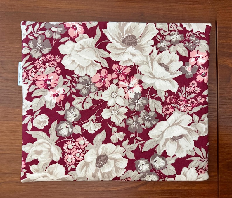 May include: A square pillow cover featuring a deep burgundy background with a dense floral pattern of large cream, light pink, and muted grey flowers and leaves. A small white fabric tag with black text is attached to the left side edge.