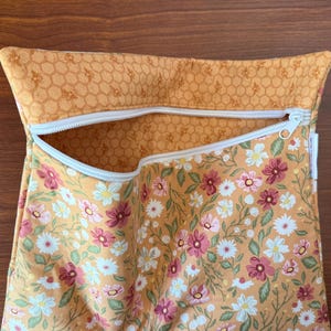 May include: A zippered pouch with a floral pattern of pink, white, and green flowers on a golden-yellow background. The top edge features a honeycomb pattern. The zipper is white.