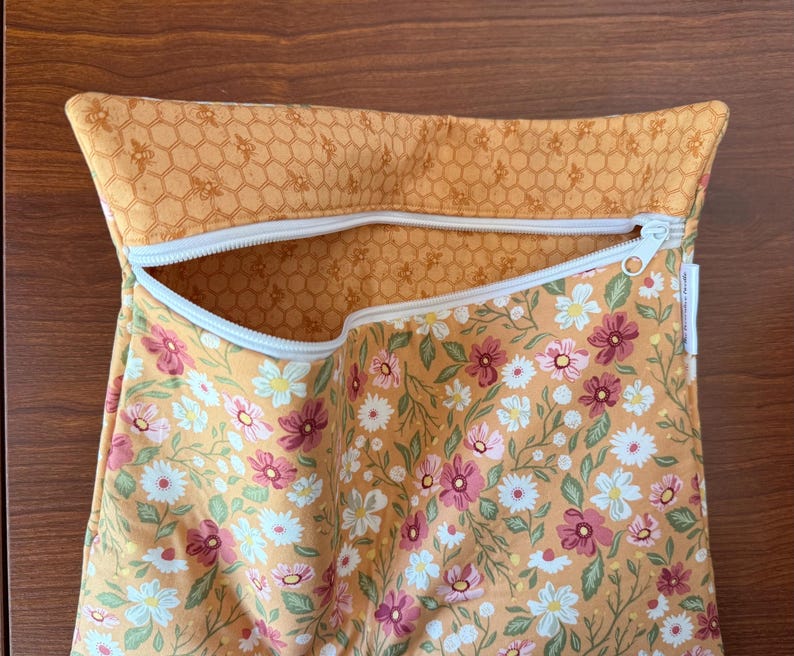 May include: A zippered pouch with a floral pattern of pink, white, and green flowers on a golden-yellow background. The top edge features a honeycomb pattern. The zipper is white.