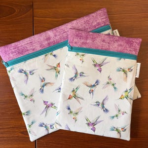 May include: Two zippered pouches with a white background and a colorful hummingbird pattern. The top of each pouch is a textured purple fabric, and the zipper is teal. The pouches are on a wooden surface.