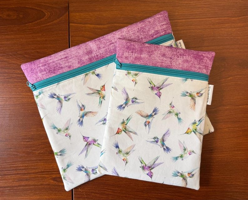 May include: Two zippered pouches with a white background and a colorful hummingbird pattern. The top of each pouch is a textured purple fabric, and the zipper is teal. The pouches are on a wooden surface.