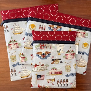 Op de afbeelding: Set van drie stoffen ritszakjes. De zakjes hebben een lichtgrijze achtergrond met patriottische illustraties van schepen, gebouwen en vlaggen. De bovenkant en de rand zijn rood met witte sterren. De ritsen zijn zwart.