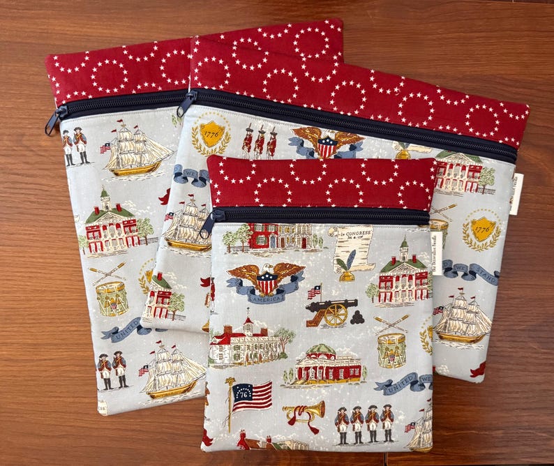 Op de afbeelding: Set van drie stoffen ritszakjes. De zakjes hebben een lichtgrijze achtergrond met patriottische illustraties van schepen, gebouwen en vlaggen. De bovenkant en de rand zijn rood met witte sterren. De ritsen zijn zwart.