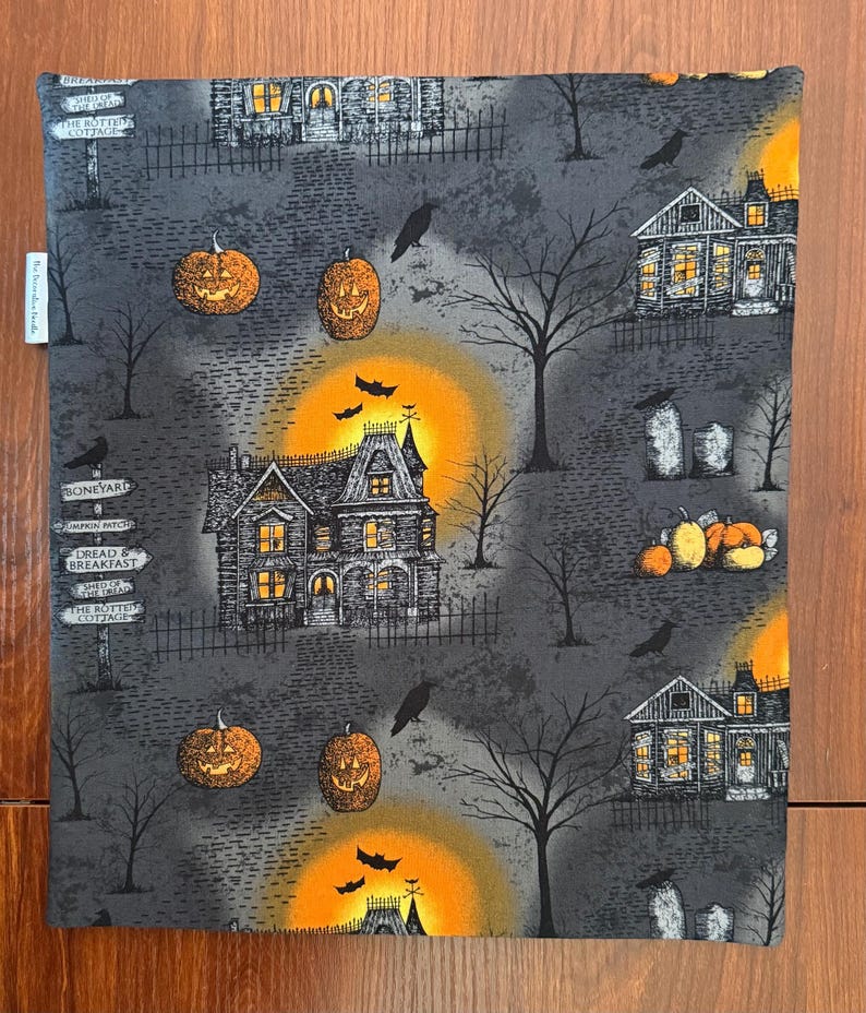 May include: A rectangular fabric panel featuring a Halloween-themed print on a dark gray background. The pattern includes haunted houses, glowing orange moons, jack-o'-lanterns, bare trees, black crows, bats, and signposts with text like Boneyard.