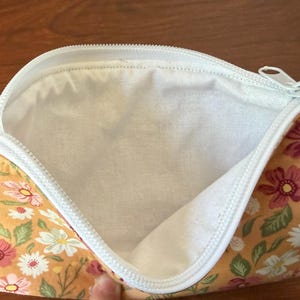 May include: Open floral print pouch with a white zipper. The pouch features a yellow background with pink, white, and green floral patterns. The interior is white.