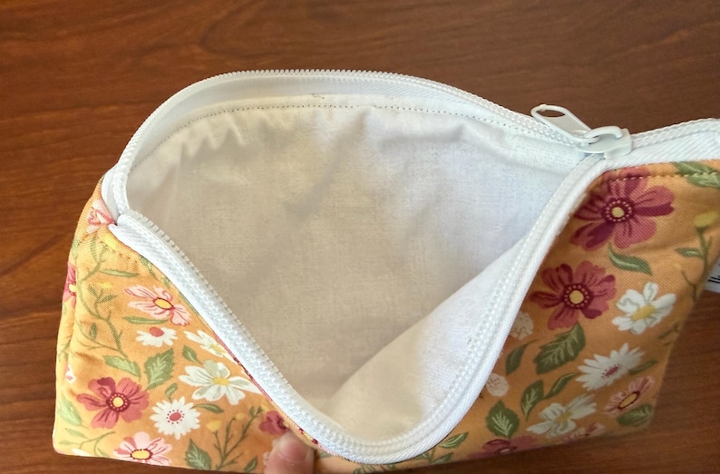 May include: Open floral print pouch with a white zipper. The pouch features a yellow background with pink, white, and green floral patterns. The interior is white.