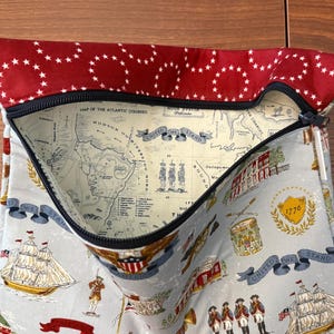 Op de afbeelding: Een stoffen etui met een rode bovenkant met witte sterren en een zwarte ritssluiting. De hoofdgedeelte van de etui is lichtblauw met historische illustraties, waaronder schepen, kaarten en de tekst "United We Stand."