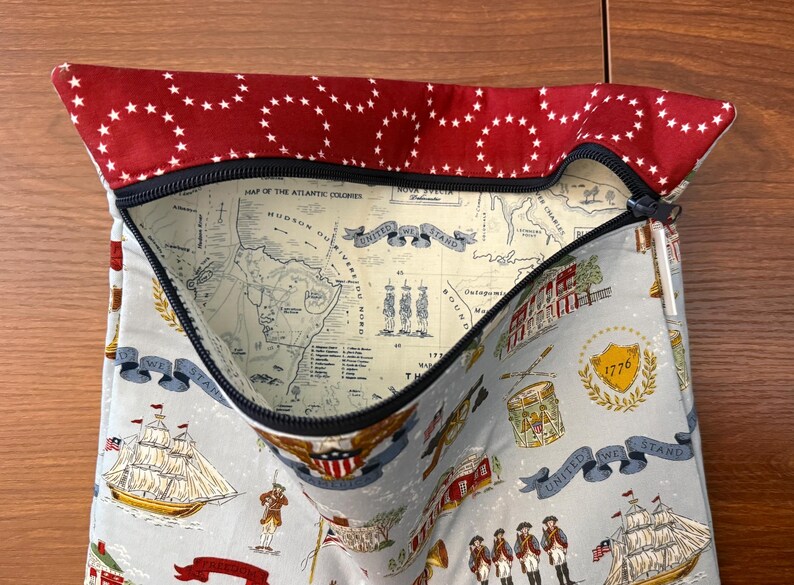 Op de afbeelding: Een stoffen etui met een rode bovenkant met witte sterren en een zwarte ritssluiting. De hoofdgedeelte van de etui is lichtblauw met historische illustraties, waaronder schepen, kaarten en de tekst "United We Stand."