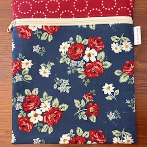 K&ouml;nnte beinhalten: Rechteckige Stofftasche mit Rei&szlig;verschluss. Der Hauptteil zeigt ein marineblaues Blumenmuster mit roten Rosen, wei&szlig;en G&auml;nsebl&uuml;mchen und gr&uuml;nen Bl&auml;ttern. Der obere Teil ist rot mit einem wei&szlig;en Sternmuster. Ein kleines wei&szlig;es Etikett ist angebracht.